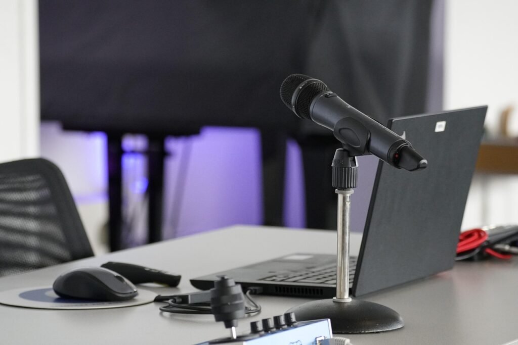 Handheld microphone on a desk stand beside a laptop, mouse and audio interface