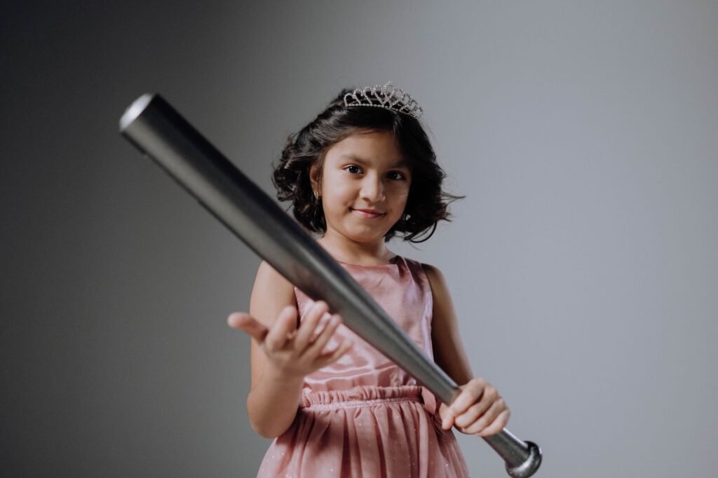 A confident young girl in a tiara holding a baseball bat against a gray background