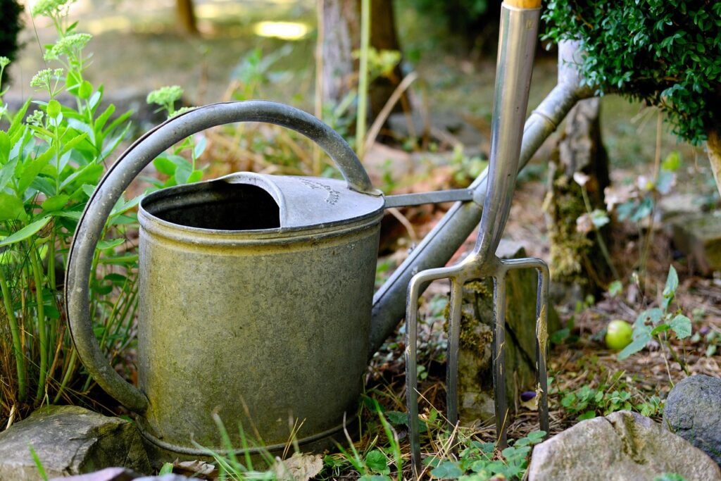 A four tine garden fork standing in the soil of a garden bed beside a watering can